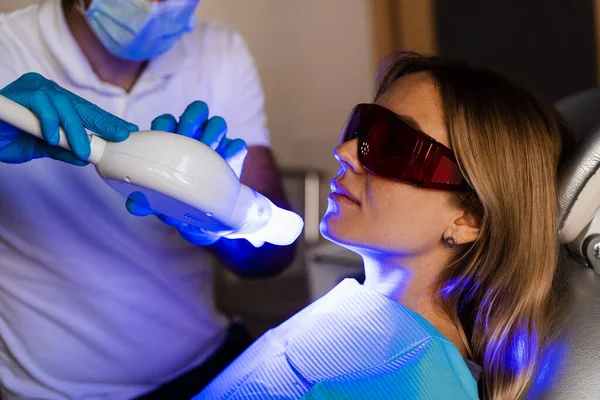 Artificial teeth whitening. Dental ultraviolet whitening treatment with light, fluoride and laser. Woman in red protective glasses patient do UV teeth whitening procedure