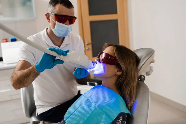 Artificial teeth whitening. Woman in red protective glasses patient do UV teeth whitening procedure. Dental ultraviolet whitening treatment with light, fluoride and laser