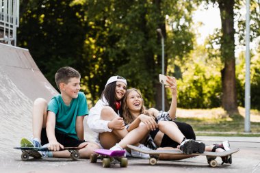 Kids smile and laugh and making selfie on phone together with skateboard and penny boards on the sports ramp playground. Children friendship concept