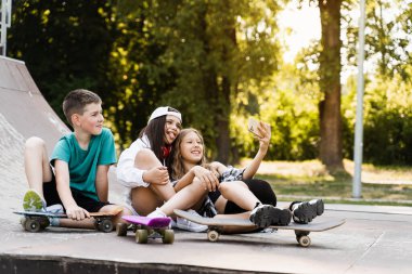 Kids smile and laugh and making selfie on phone together with skateboard and penny boards on the sports ramp playground. Children friendship concept