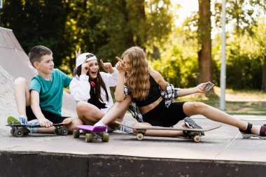 Kids smile and laugh and making selfie on phone together with skateboard and penny boards on the sports ramp playground. Children friendship concept