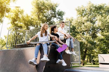 Sports kids with skateboard and penny boards are sitting and chatting with each other on sports ramp on playground. Children communication and friendship. Sports extreme lifestyle