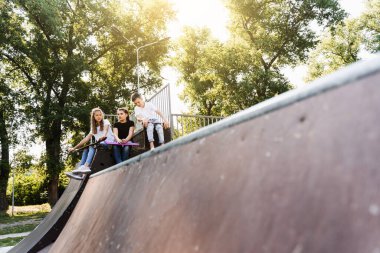 Sports kids with skateboard and penny boards are sitting and chatting with each other on sports ramp on playground. Children communication and friendship. Sports extreme lifestyle