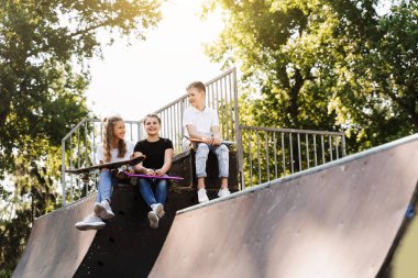 Sports kids with skateboard and penny boards are sitting and chatting with each other on sports ramp on playground. Children communication and friendship. Sports extreme lifestyle