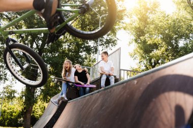 Bmx bicycle rider making tricks and sports kids with skateboard and penny boards are sitting and chatting on background. Children communication and friendship on sports ramp on playground