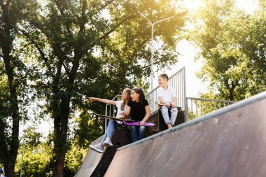 Sports kids with skateboard and penny boards are sitting and chatting with each other on sports ramp on playground. Children communication and friendship. Sports extreme lifestyle