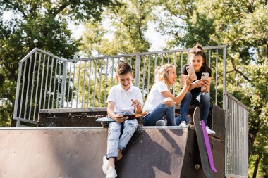 Phone addicted sports children with skateboard and penny boards are sitting and looking at smartphones on sports ramp on playground. Kids addiction of phones