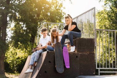 Phone addicted sports children with skateboard and penny boards are sitting and looking at smartphones on sports ramp on playground. Kids addiction of phones