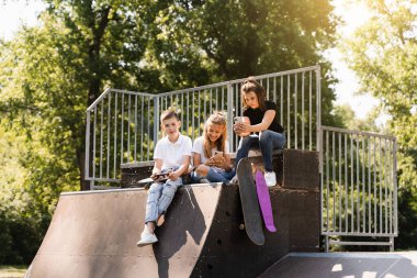 Phone addicted sports children with skateboard and penny boards use phones instead of skating and play together. Children looking at smartphones on sports ramp. Kids addiction of phones