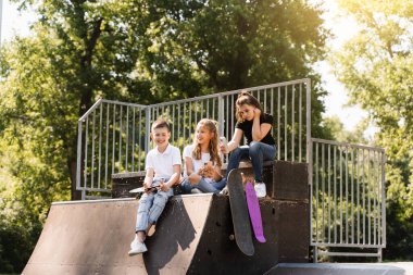 Phone addicted sports children with skateboard and penny boards use phones instead of skating and play together. Children looking at smartphones on sports ramp. Kids addiction of phones