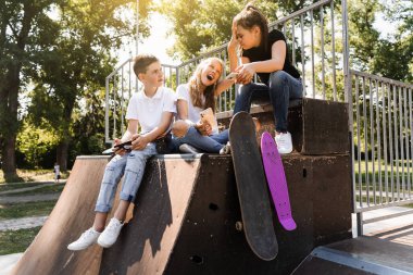 Phone addicted sports children with skateboard and penny boards smiling and looking at smartphones on sports ramp on playground. Kids addiction of phones