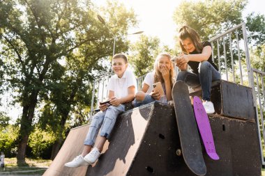 Phone addicted sports children with skateboard and penny boards smiling and looking at smartphones on sports ramp on playground. Kids addiction of phones