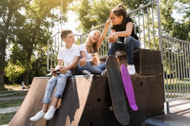Phone addicted sports children with skateboard and penny boards smiling and looking at smartphones on sports ramp on playground. Kids addiction of phones