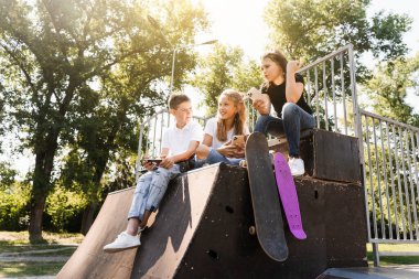 Phone addicted sports children with skateboard and penny boards smiling and looking at smartphones on sports ramp on playground. Kids addiction of phones
