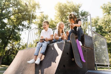 Phone addicted sports children with skateboard and penny boards smiling and looking at smartphones on sports ramp on playground. Kids addiction of phones