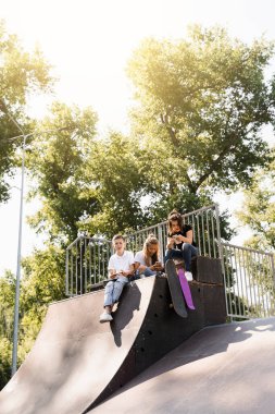 Phone addicted sports children with skateboard and penny boards use phones instead of skating and play together. Children looking at smartphones on sports ramp. Kids addiction of phones