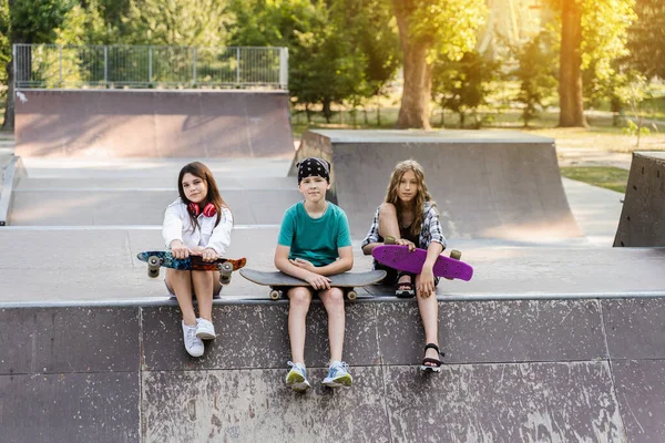 Children friends company with skateboard and penny boards on skate board park. Extreme lifestyle. Active kids sitting on sport ramp, communicate with each other and have fun