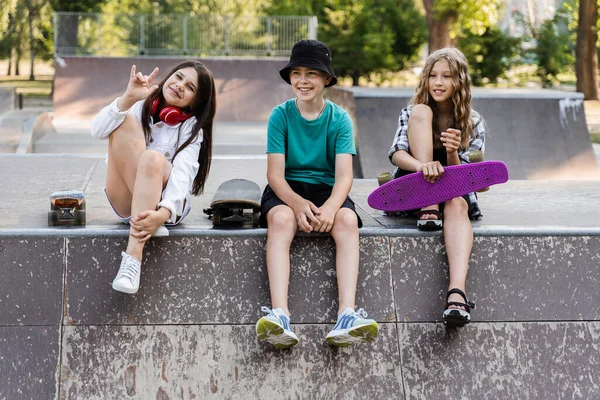 Children friends company with skateboard and penny boards on skate board park. Extreme lifestyle. Active kids sitting on sport ramp, communicate with each other and have fun