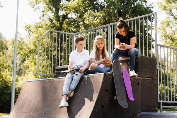 Phone addicted sports kids with skateboard and penny boards are sitting and looking at smartphones on sports ramp on playground. Children addiction of phones