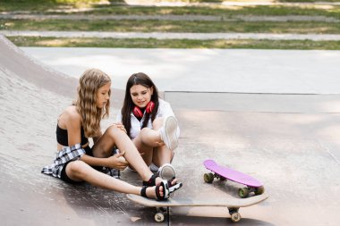 Falling from skateboard. Active children girls after fall from penny board injured, sitting and showing bruise each others on sport ramp on skate park playground