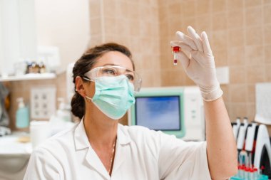 Laboratuvar asistanı elinde kanla test tüpünü tutuyor ve bakıyor. Laboratuvarda kan testi. Gözlüklü ve maskeli tıp bilimcisi