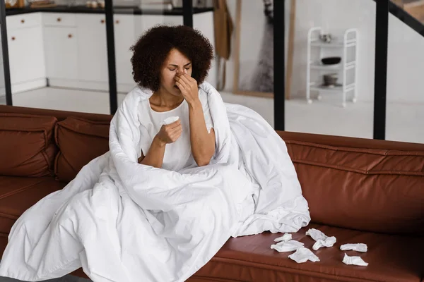 病気のアフリカの少女インフルエンザは 自宅でソファに座っている間 彼女の鼻を吹いてナプキンにくしゃみをします 寒さと毛布に包まれた女性の鼻水 — ストック写真