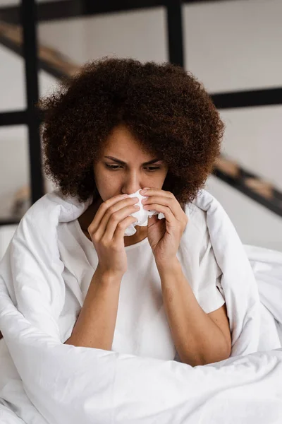 Runny nose african girl flu blows her nose and sneezes into napkin while sitting sofa at home. Fever and cold of woman wrapped in blanket