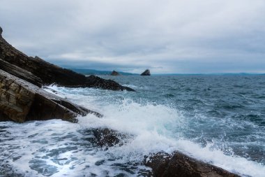 Arka planda ufuk çizgisi olan deniz ve kayalar. Bulutlu ve yağmurlu, dalgalı.