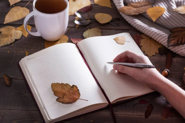 秋を書く女性の手は黄色の葉とお茶のカップとノートブックにリストを行いたいです 茶色の木製の背景にスペースをコピー ストック写真