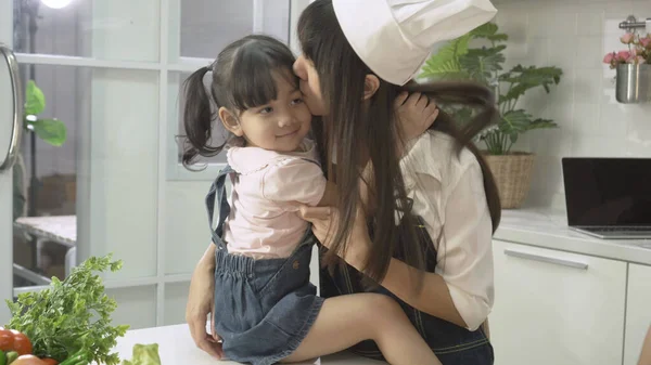 happy family, Asian Mother hug and kiss cheek of her cute little daughter while cooking in kitchen. children girls playful learning to assistant cook. kids and mom enjoy preparing meal in house.