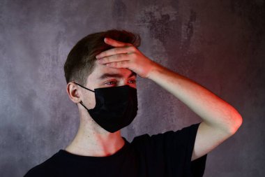 Koruyucu tıbbi maske takmış yakışıklı bir çocuk başını tutuyor, başı ağrıyor.