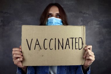 Elinde aşı posteri olan tıbbi koruyucu maskeli genç bir kadın.