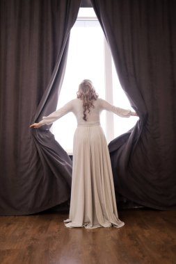 Güzel, uzun elbiseli genç bir kadın pencere perdelerini açıyor, arkadan bir manzara. Dikey yönelim