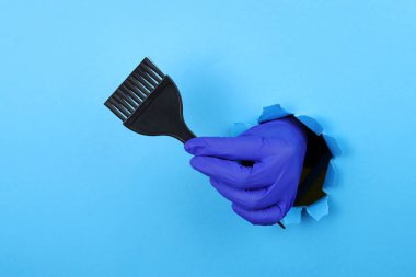 Kuaförün eli, mavi kağıttaki yırtık bir deliğin içinden saç fırçasını tutuyor. Saç boyası