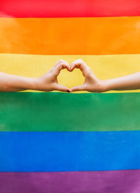 Arka planda LGBTQ gökkuşağı bayrağı olan bir kalp oluşturan iki kadının ellerini kapatın. Homoseksüeller için gurur ayı ve cinsel ve aşk çeşitliliğinin kutlandığı gün.