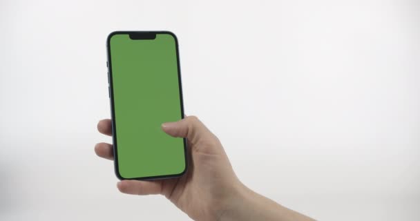 女性は左に緑の画面垂直とスワイプ写真や写真とスマートフォンを保持しています 白の背景に緑の画面でスマートフォン クロマキーモックアップオンスマートフォンインハンド — ストック動画