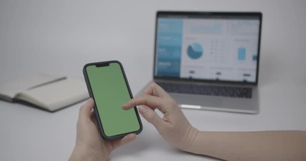 クロマキー手にスマートフォンでモックアップ緑の画面で電話を使用する女性 画面上のチャートと背景にメモ帳付きノートパソコン — ストック動画