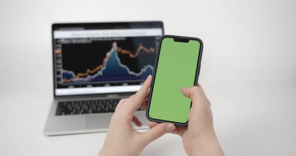クロマキー手にスマートフォンでモックアップ緑の画面で電話を使用する女性 画面上のチャートと背景にメモ帳付きノートパソコン — ストック動画