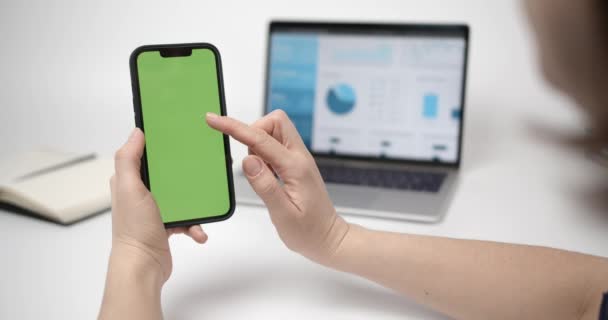 クロマキー手にスマートフォンでモックアップ緑の画面で電話を使用する女性 画面上のチャートと背景にメモ帳付きノートパソコン — ストック動画