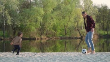 Baba, Oğul 'la Kum' da Futbol oynuyor. Ağır çekim. Genç adam ve küçük çocuk parkta siyah ve beyaz futbol topuyla oynuyor.