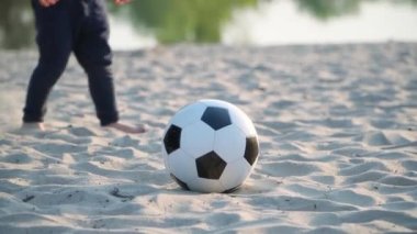 Küçük çocuk futbol topunu tekmeliyor. Baba ve Oğul, nehir kenarında siyah ve beyaz futbol topuyla oynuyor. 4k