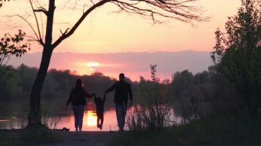 Parents Hold Kid by Hands and Walk on Banks of River against Background of Beautiful Sunset. Slow Motion