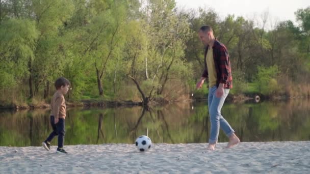 父亲和儿子在沙地上踢足球慢动作 年轻人和小男孩在河边附近的室外玩黑白足球 — 图库视频影像
