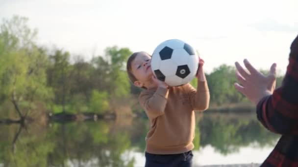 小男孩接球和扔球给他的父亲 男子和儿童在河附近的室外玩黑白足球 — 图库视频影像
