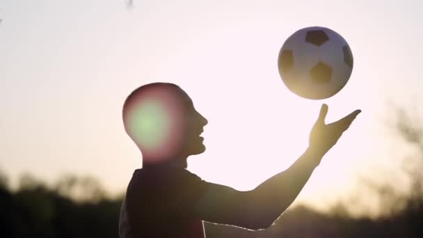 男は彼の頭でサッカーボールをキック若い男のシルエットは空の背景に彼の頭でサッカーボールをキックし 太陽スローモーションを設定します — ストック動画