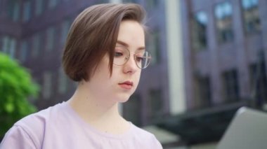 Gözlüklü genç kız dizüstü bilgisayara bakar sonra yüzünü kameraya çevirir ve gülümser. Kız Dışarda Dizüstü bilgisayar Kullanıyor