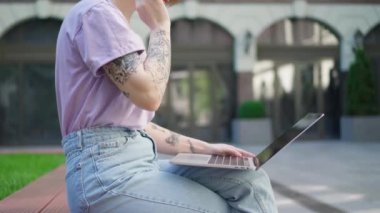 Diz üstü bilgisayarı dışarıda oturan dövmeli genç kız. Kız öğrenci ya da bilişim uzmanı şehir merkezindeki bankta oturan dizüstünü kullanıyor