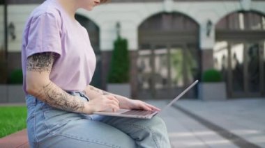Diz üstü bilgisayarı dışarıda oturan dövmeli genç kız. Güzel kız, modern ofis binasının avlusunda otururken bilgisayarını uzaktan çalışma için kullanıyor.