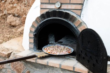 Pizzayı el yapımı beyaz odun fırınına koyan adam açık havada kürek ve arka planda yapılmış.
