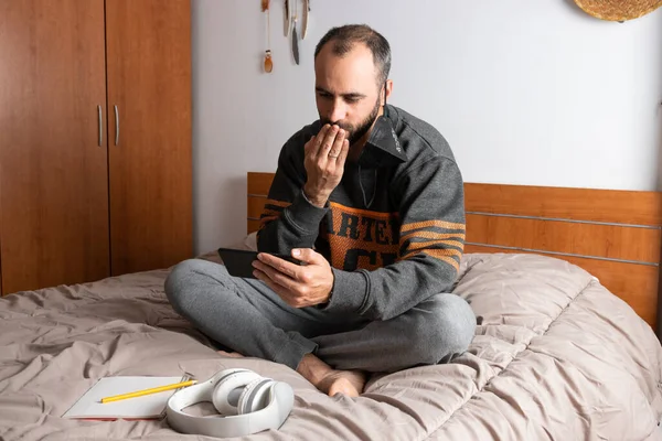 Человек в наушниках, мобильном телефоне и маске, сидящий на кровати и запертый в своей комнате — стоковое фото